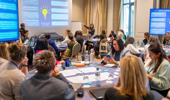 Women participating in a leadership workshop discuss activities around tables while a facilitator presents different leadership styles at the front of the room.